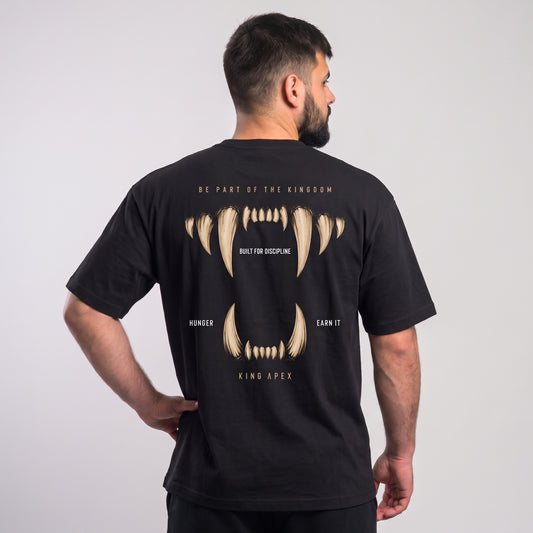 Male model wearing the black KING APEX Hunger oversized T-shirt, back view with one hand on the hip showing the gold fangs framing BUILT FOR DISCIPLINE, HUNGER and EARN IT lettering.