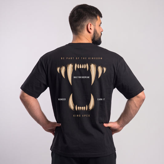 Male model wearing the black KING APEX Hunger oversized T-shirt, back view with hands on hips showing the gold fangs framing BUILT FOR DISCIPLINE, HUNGER and EARN IT lettering.