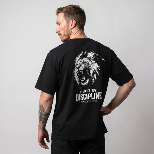 Male model wearing the black KING APEX Lion Built by Discipline oversized T-shirt, back view with one hand on the hip showing the white roaring lion portrait with BUILT BY DISCIPLINE lettering.