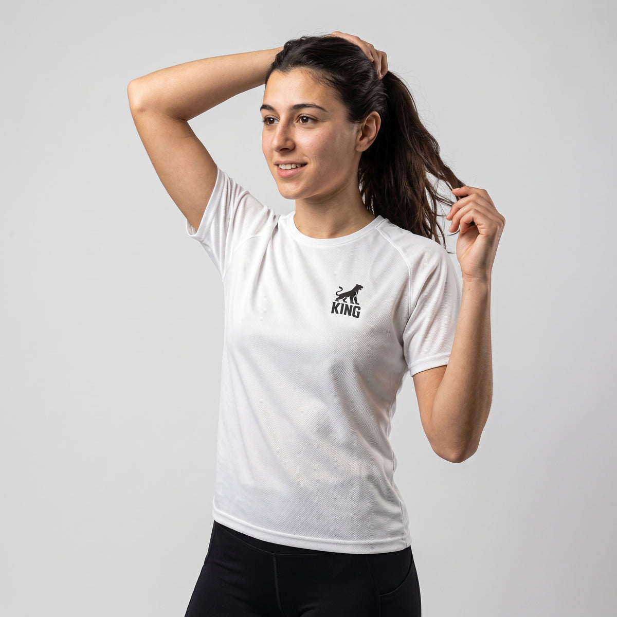 Three-quarter front view of woman wearing the white KING APEX Lioness sports jersey while adjusting her hair.