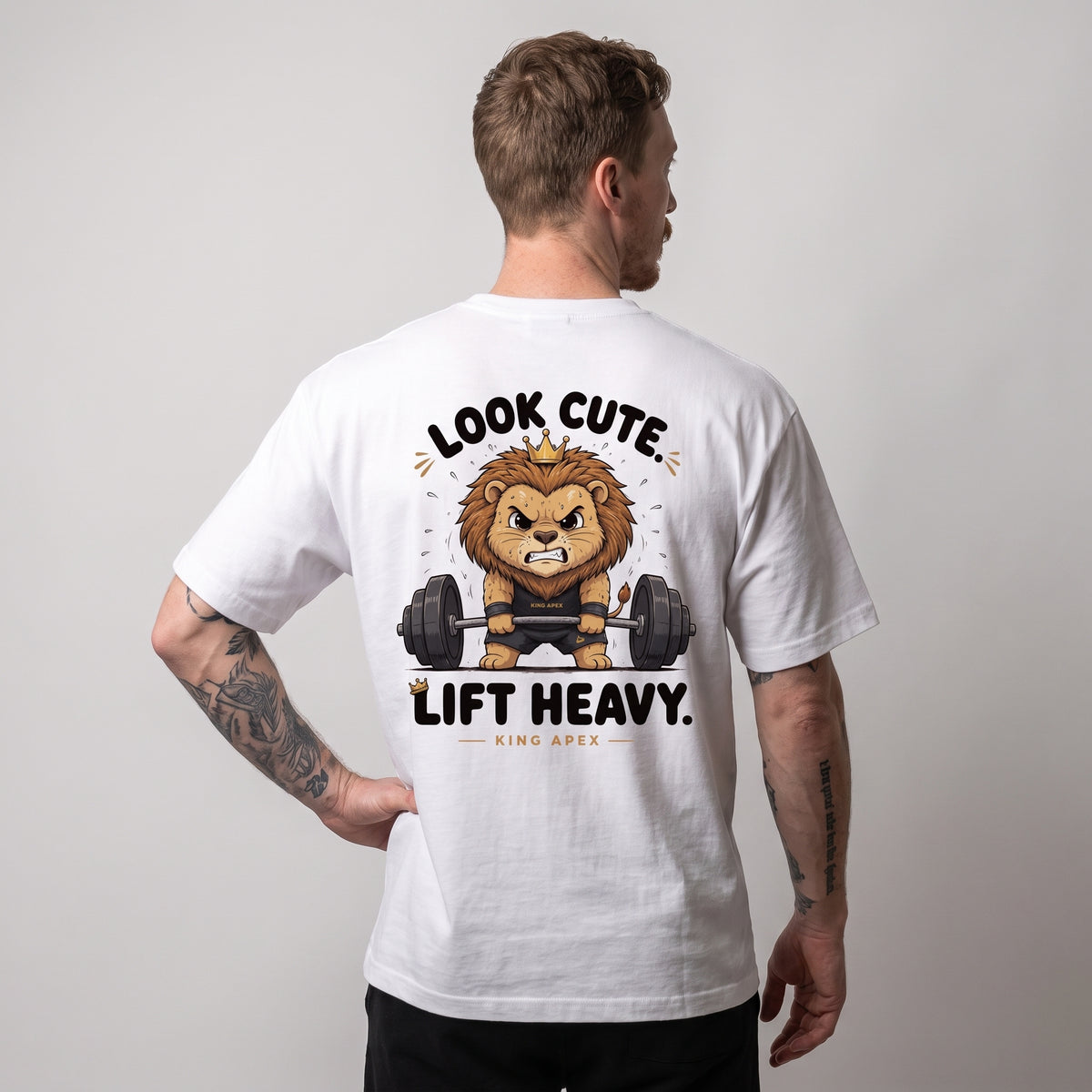 Male model wearing the white KING APEX Look Cute. Lift Heavy. oversized T-shirt, back view with one hand on the hip showing the crowned cartoon lion lifting a barbell with LOOK CUTE. LIFT HEAVY. lettering.