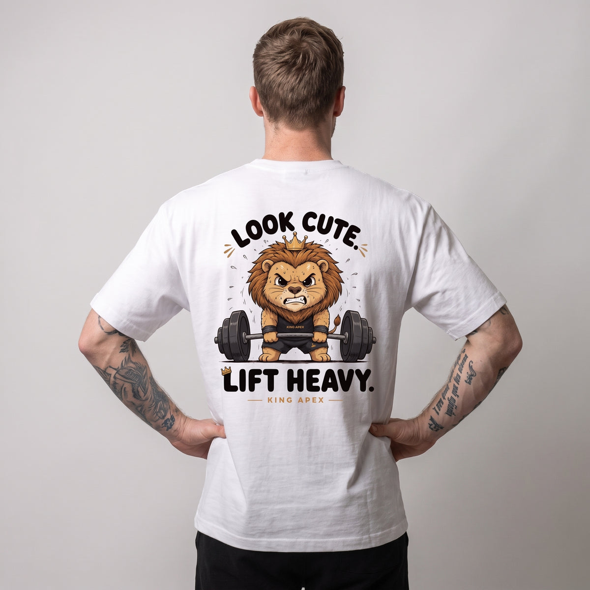 Male model wearing the white KING APEX Look Cute. Lift Heavy. oversized T-shirt, back view with hands on hips showing the crowned cartoon lion lifting a barbell with LOOK CUTE. LIFT HEAVY. lettering.