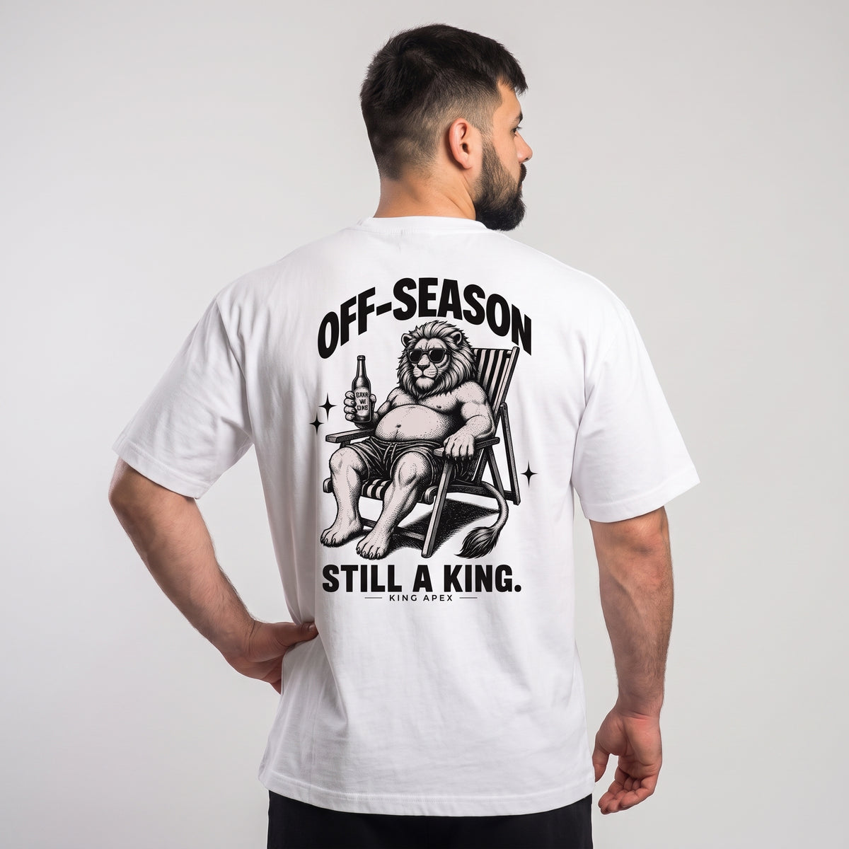 Male model wearing the white KING APEX Off-Season oversized T-shirt, back view with one hand on the hip showing the black reclining lion artwork with OFF-SEASON and STILL A KING. lettering.