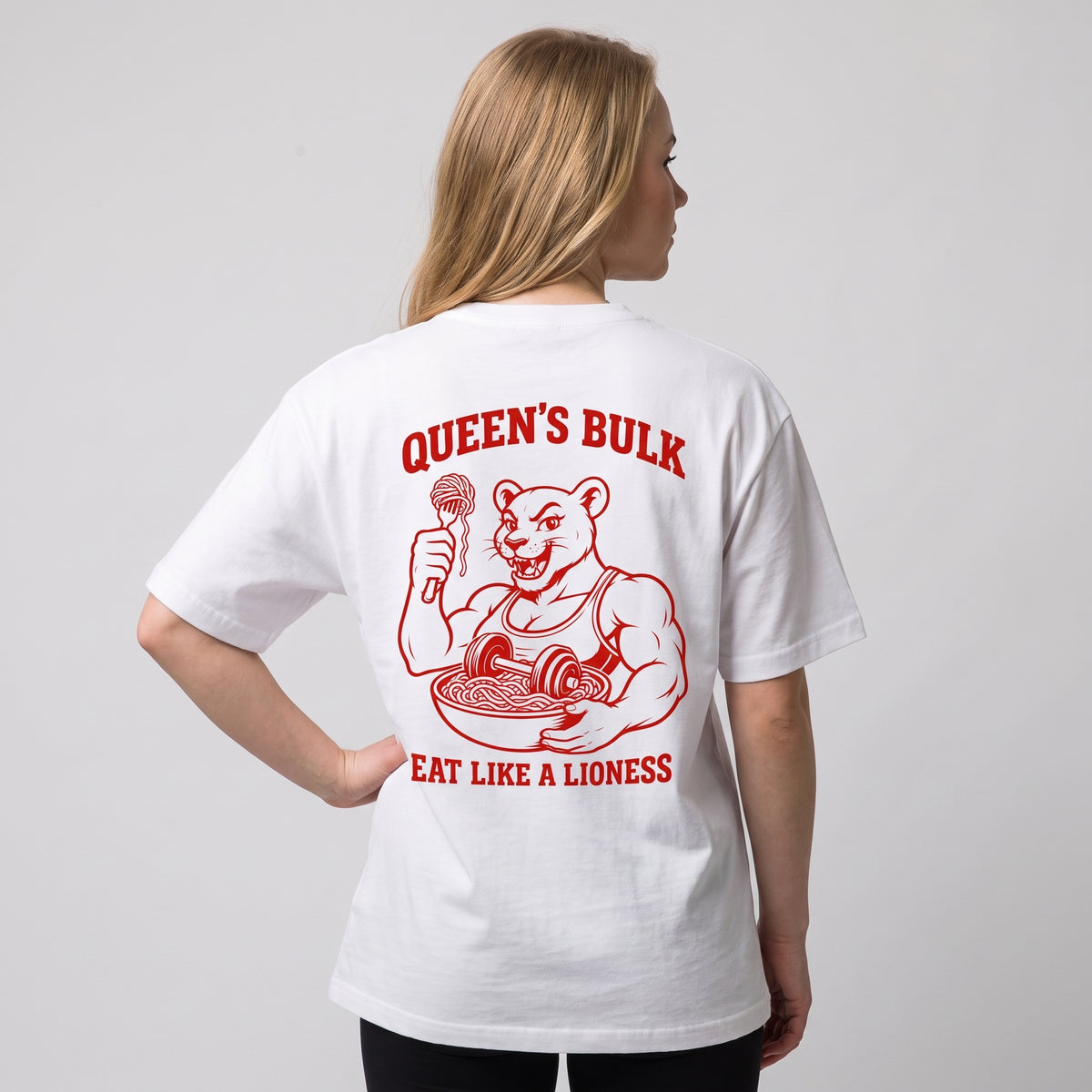 Female model wearing the white KING APEX Queen’s Bulk oversized T-shirt, back view with one hand on the hip showing the red lioness eating pasta with QUEEN’S BULK and EAT LIKE A LIONESS lettering.