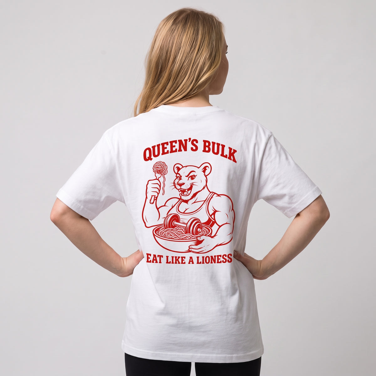 Female model wearing the white KING APEX Queen’s Bulk oversized T-shirt, back view with hands on hips showing the red lioness eating pasta with QUEEN’S BULK and EAT LIKE A LIONESS lettering.